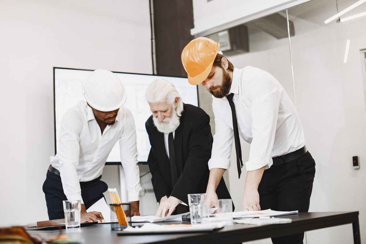 why-choose-us-02 Group of architects discussing construction blueprints wearing hardhats in a modern office.