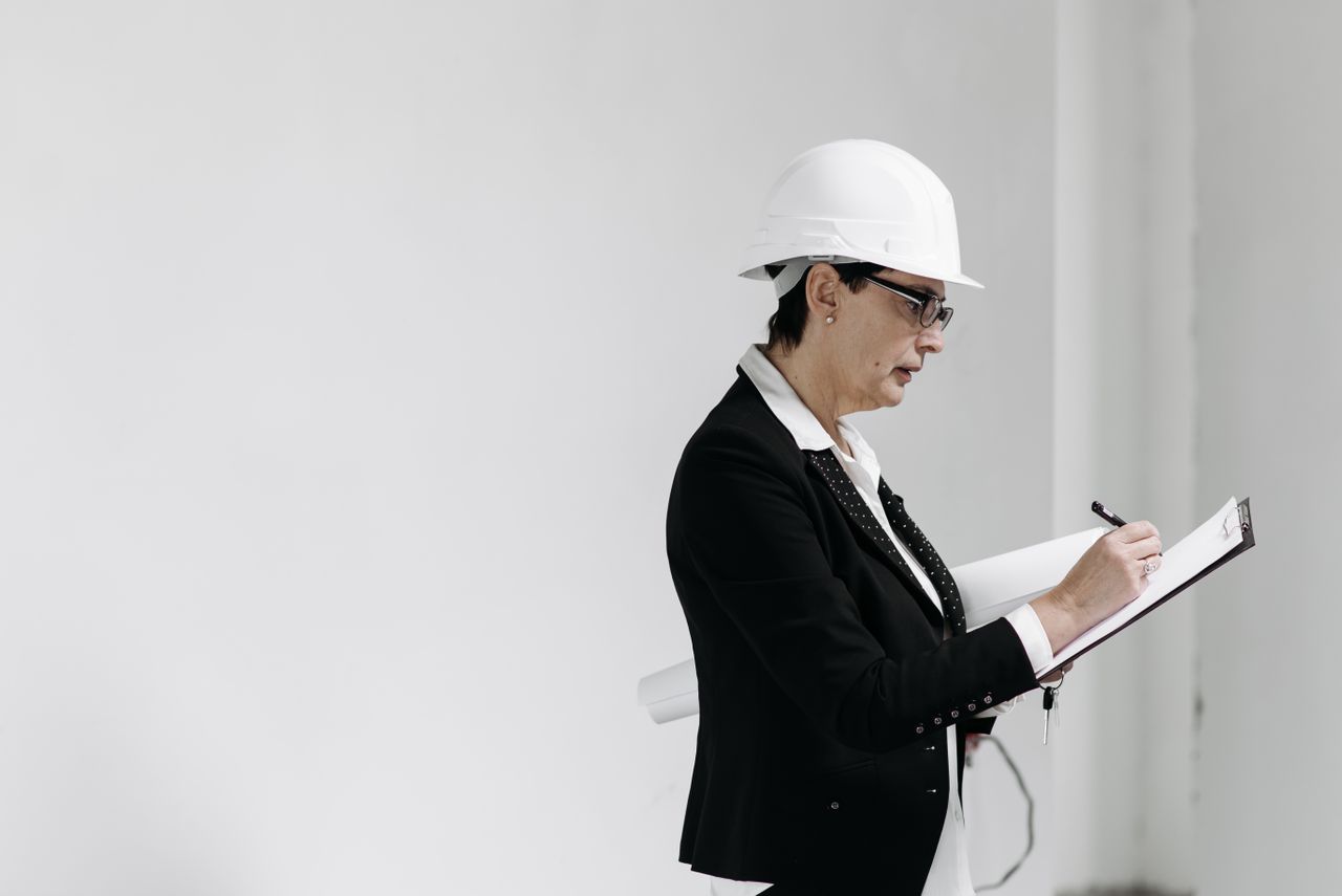 why-choose-us-01 Woman in business attire with hard hat taking notes at a construction site.